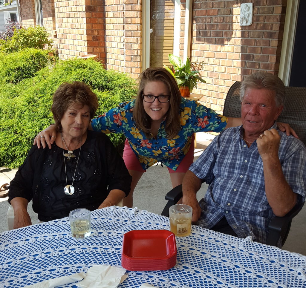 Ashlyne is wearing a bright floral shirt and pink shorts, putting her arms around her Gramma and Papa at her 30th birthday party. Her grandparents are the best.