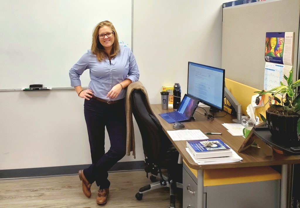 Ashlyne stands near her desk, leaning on her chair wearing a blue button-up, navy pants, and a smile. This is her office in Open Learning.