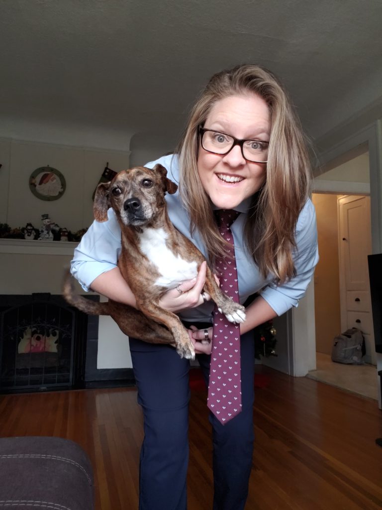 Ashlyne leans in toward the camera holder her dog who has a concerned look on her face. Ashlyne is smiling and wearing a burgundy tie, light blue shirt, and navy pants.