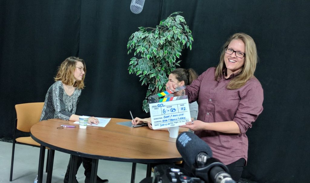 Ashlyne laughs while holding a clapper because she is excited about producing educational videos. Two actresses sit behind her rehearsing their lines at a round table.