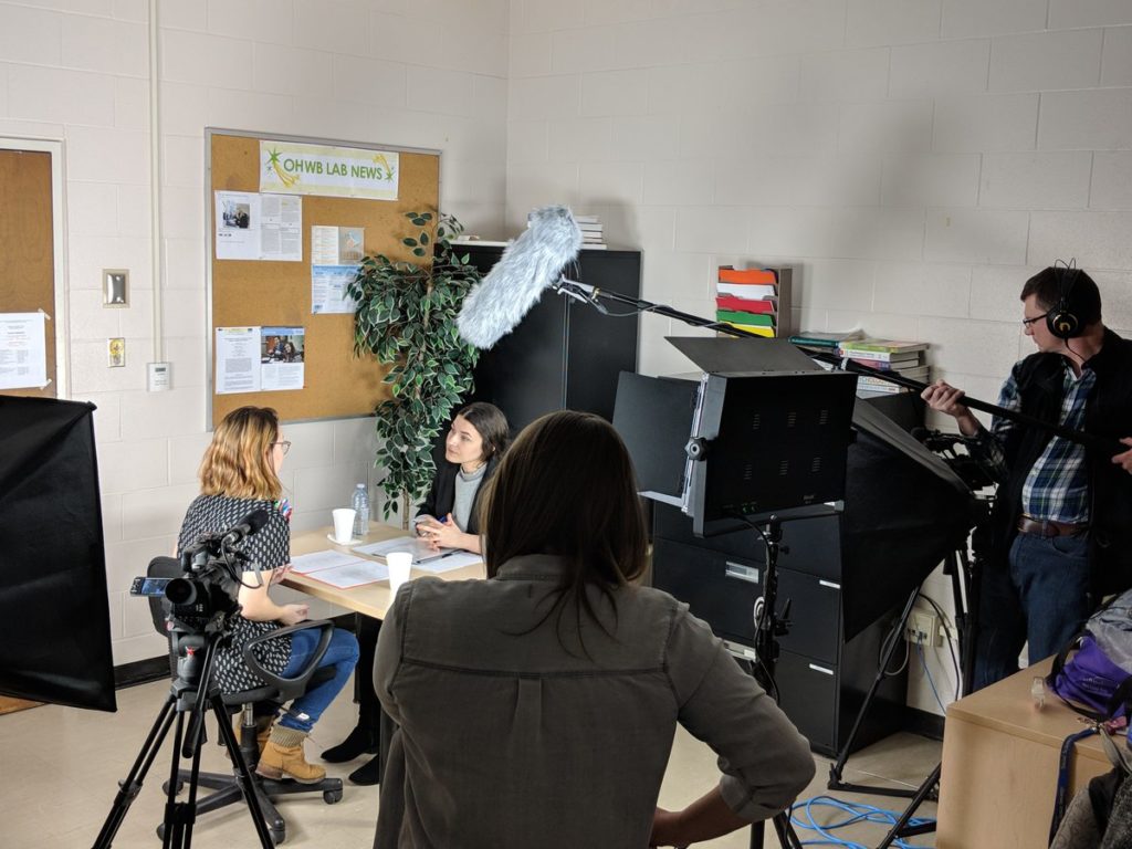 A light shines on two actors sitting at a table with a boom mic above them. Ashlyne stands darkened in the foreground with her back to the camera, pondering director's notes