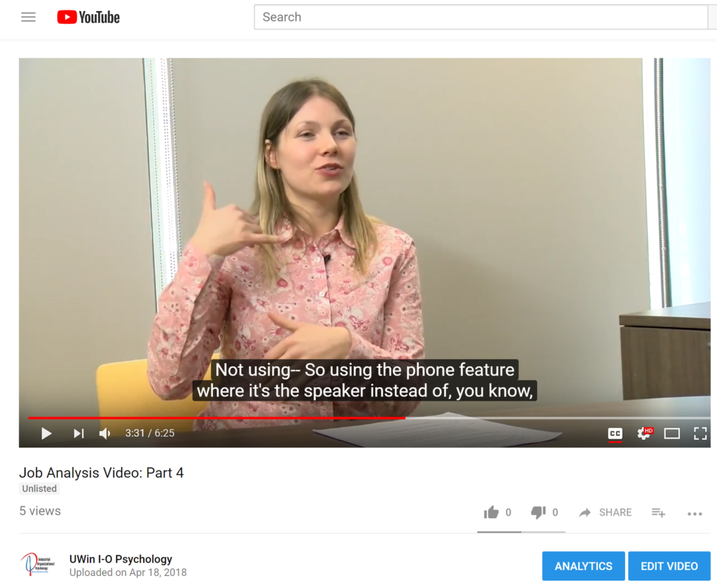 A woman sits behind a table wearing a pink blouse, making a phone sign with her pinky finger and thumb as she describes her job duties as part of a video about job analysis. This is a screenshot.