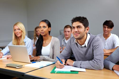 Students sitting at a desk taking notes. One student is looking to the front of the room and using a laptop to type her notes, while the student to her right is using paper and pen to hand write his notes. 