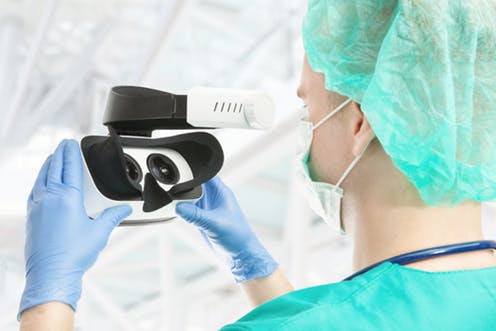 Photo of a medical student in scrubs, hair net, gloves and a mask holding virtual reality glasses in front of their face. 